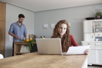 Femme à la maison en utilisant un ordinateur portable avec l'homme en arrière-plan coupe de légumes — Photo de stock