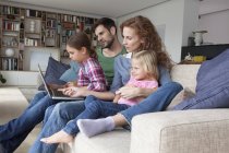 Coppia seduta con loro con due figlie piccole sul divano in soggiorno guardando il computer portatile — Foto stock