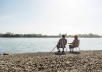 Rückansicht eines älteren Ehepaares, das nebeneinander auf Klappstühlen am Flussufer sitzt — Stockfoto