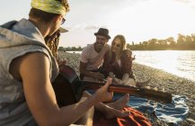 Amigos relajándose a orillas del río con guitarra al atardecer - foto de stock