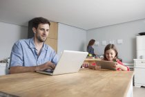 Человек и его маленькая дочь сидят за кухонным столом с помощью ноутбука и цифрового планшета — стоковое фото