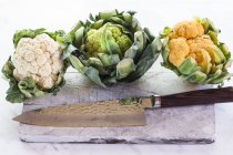 Green, orange and white cauliflowers with knife on chopping board — Stock Photo