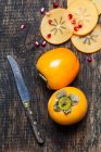 Whole and sliced kaki with knife and pomegranate seeds on dark wood — Stock Photo