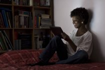 Mujer joven sentada en su cama en casa usando tableta digital - foto de stock