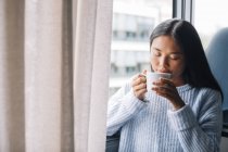 Jeune femme avec une tasse de café debout devant la fenêtre ouverte — Photo de stock