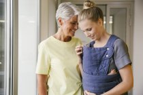 Young mother with baby carrier showing her baby to mature woman — Stock Photo