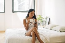 Young woman sitting on bed with cell phone and cup of coffee — Stock Photo