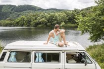 Hombre sentado en el techo de una furgoneta a orillas del lago - foto de stock