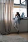 Junge Frau mit Tasse Kaffee steht vor offenem Fenster — Stockfoto
