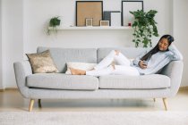 Mujer joven que se relaja en el sofá en la sala de estar con teléfono inteligente - foto de stock