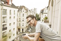 Sonriente hombre en balcón mirando por la calle sosteniendo la taza de café - foto de stock