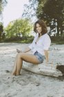 Ritratto di donna sorridente con smartphone seduta su legno morto in spiaggia — Foto stock