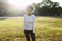 Mujer joven sonriente de pie en un prado de un parque a la luz de fondo - foto de stock