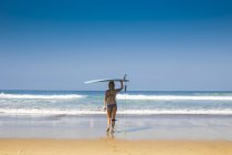 Indonesien, Bali, Rückenansicht einer Frau, die am Strand ihr Surfbrett auf dem Kopf trägt — Stockfoto