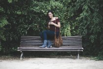 Sorrindo jovem mulher sentada em um banco no parque usando telefone celular — Fotografia de Stock