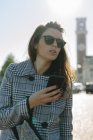 Donna che indossa cappotto a scacchi e occhiali da sole che tengono il telefono cellulare — Foto stock
