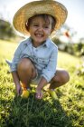 Retrato de una niña sonriente con sombrero de paja agachada en un prado - foto de stock