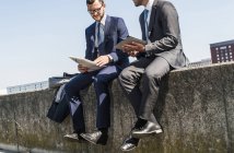 Zwei junge Geschäftsleute sitzen an einer Wand am Fluss und arbeiten — Stockfoto