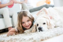 Kleines Mädchen kuschelt mit Hund, liegt auf dem Boden, Eltern zu Hause im Hintergrund — Stockfoto