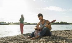 Jeune homme jouant de la guitare avec une femme au bord de la rivière — Photo de stock