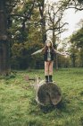 Menina vestindo boné de pé com braços estendidos em um tronco de árvore no parque outonal — Fotografia de Stock