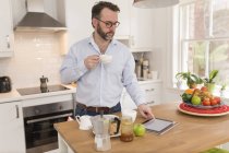 Hombre con taza de café de pie en la cocina usando tableta digital - foto de stock