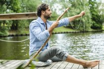 Man sitting on platform at the waterside with selfmade fishing rod — Stock Photo