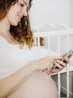 Schwangere mit Smartphone vor dem Kinderbett — Stockfoto