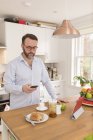 Hombre de pie en su cocina por la mañana mirando su teléfono inteligente - foto de stock