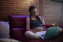 Donna incinta che utilizza il computer portatile a casa, mangiare frutta — Foto stock