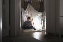 Ragazzo seduto in tenda self-made a casa la sera utilizzando tablet digitale — Foto stock