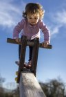 Toddler girl standing on wooden rocker against blue sky on playground — Stock Photo