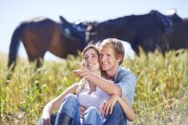 Pareja joven sentada en el campo con caballos de fondo - foto de stock