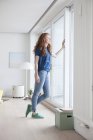 Jeune femme debout dans son salon regardant par la fenêtre — Photo de stock