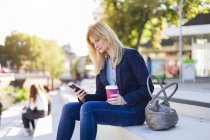 Блондинка бізнес-леді з кавою, щоб піти дивитися на смартфон — стокове фото