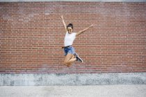 Young woman jumping up with hands up, brick wall on background — Stock Photo