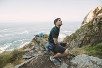 Альпинист сидит на скале у берега и смотрит вверх — стоковое фото
