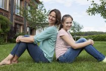 Smiling mother and daughter sitting back to back in garden — Stock Photo
