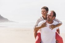 Happy mature man carrying wife piggyback on the beach — Stock Photo