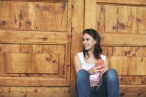 Smiling young woman hearing music with ear phones in front of a wooden door — Stock Photo