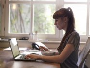 Femme au bureau utilisant un ordinateur portable et mangeant du muesli — Photo de stock