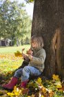 Niña apoyada en el tronco del árbol mirando un montón de hojas de otoño en sus manos - foto de stock