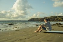 Adolescente sentado en la playa viendo atardecer - foto de stock