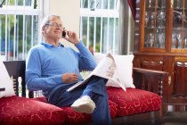 Uomo anziano al telefono che legge il giornale a casa — Foto stock
