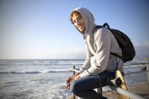 Smiling young man sitting on railing at the coast — Stock Photo