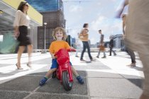 Boy riding plastic tricycle between crowd of people in city — Stock Photo