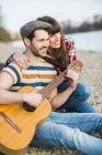 Couple sitting by the riverside, playing guitar — Stock Photo