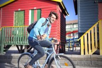 Joven sonriente montando en bicicleta en coloridas cabañas de playa - foto de stock
