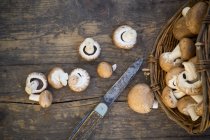 Fresh brown button mushrooms with basket  and knife on dark wood — Stock Photo