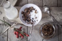 Bowl with granola with baked oats, nuts and raisins, bottle of milk and rose hips on wood — Stock Photo